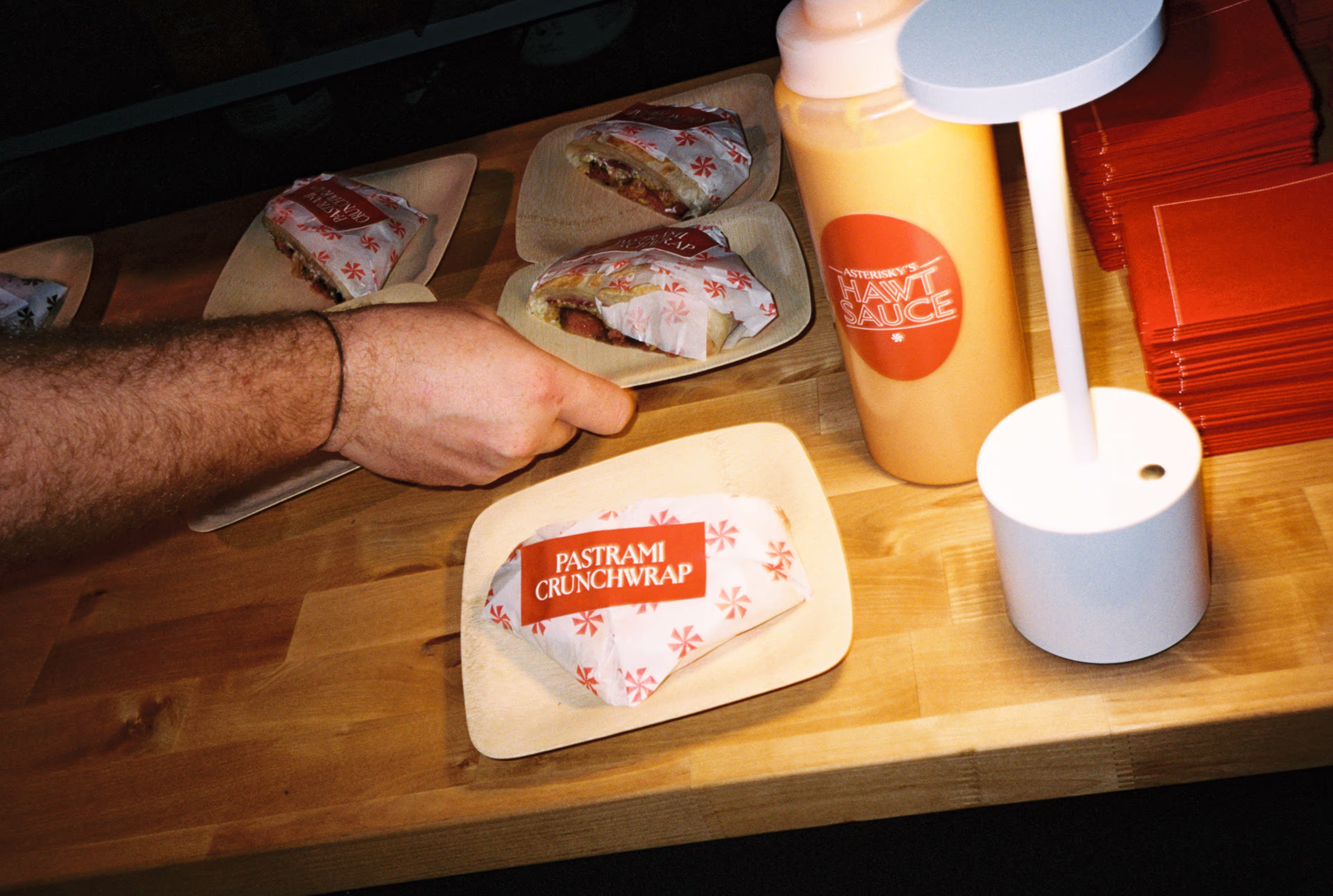 Hand grabbing a Pastrami Crunchwrap wrapped in branded paper on a wooden plate.