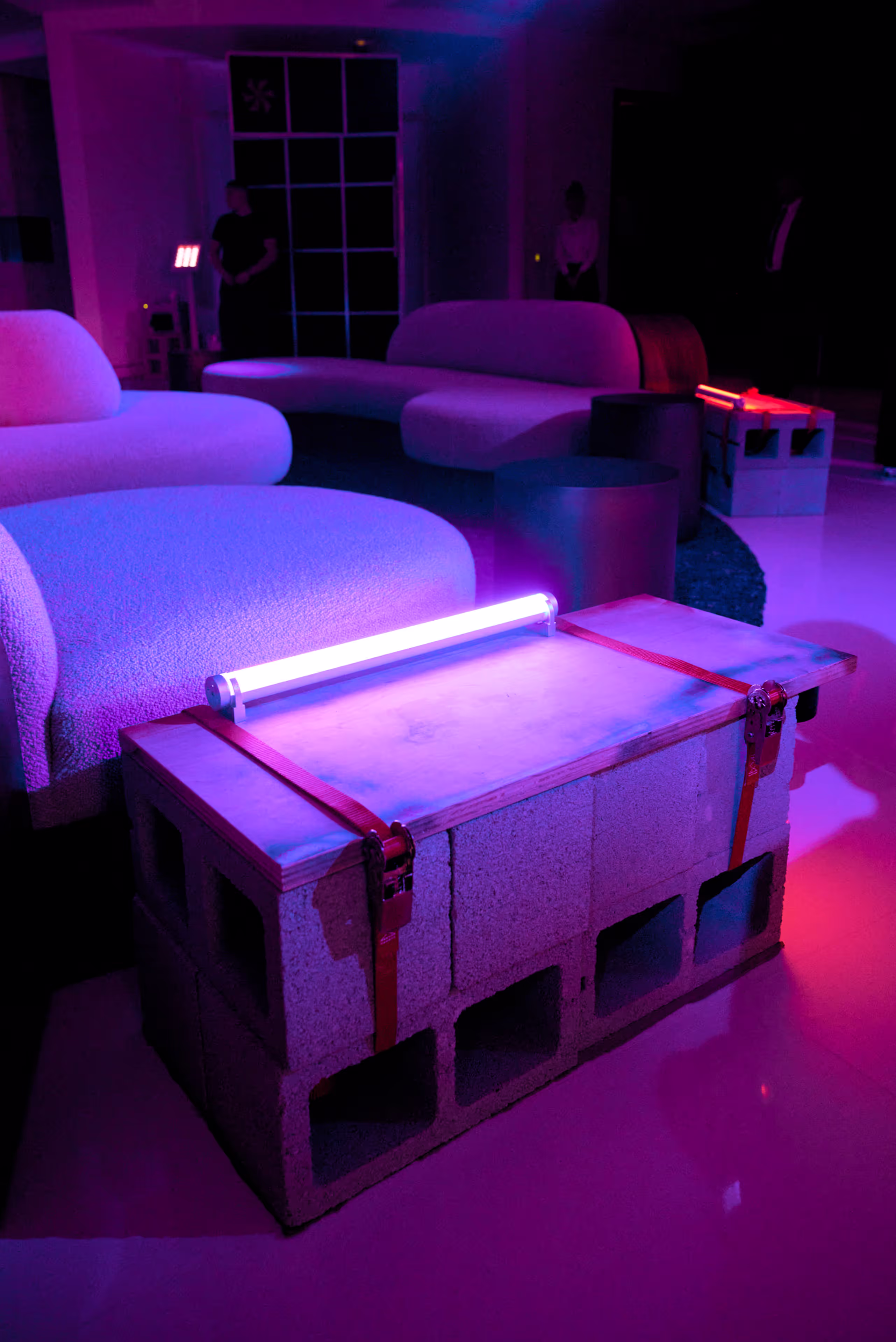 Modern seating area with curved sofas and a cinder block coffee table topped with wooden planks and a glowing neon tube light in purple lighting.