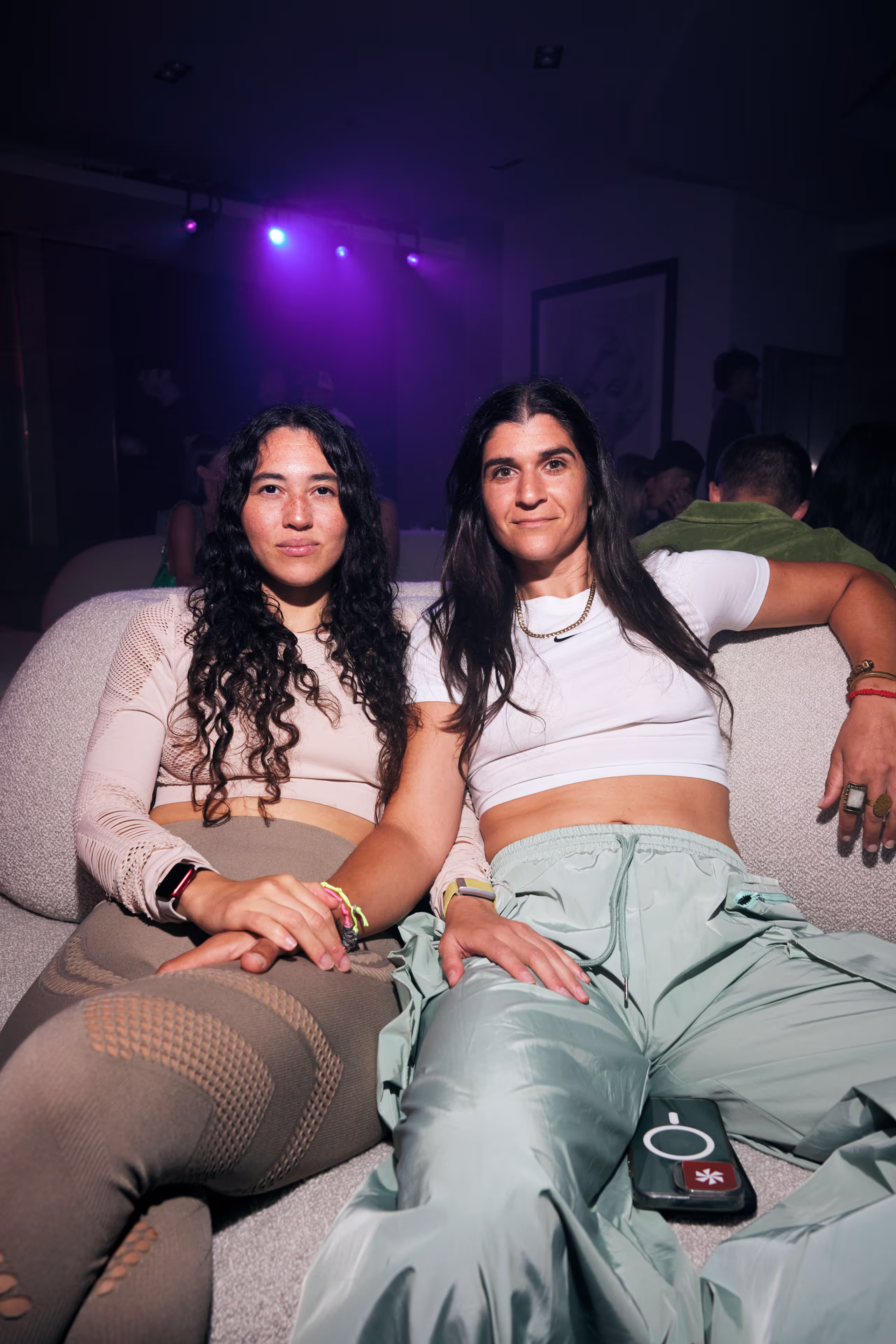 Two women sitting closely on a couch in a dimly lit room with purple lights in the background.