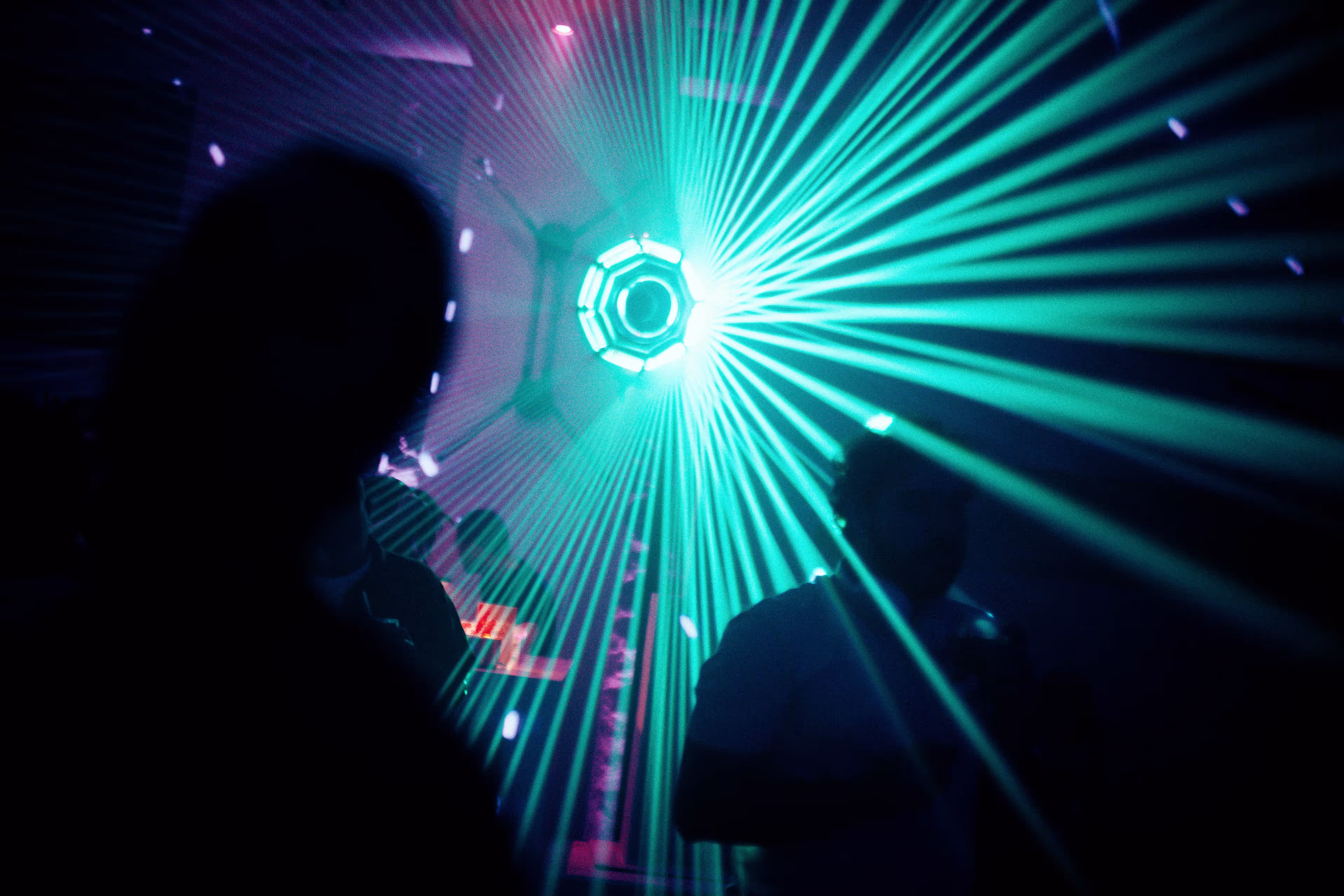 Silhouettes of people in a dark room with green laser beams radiating from a central light source on the ceiling.