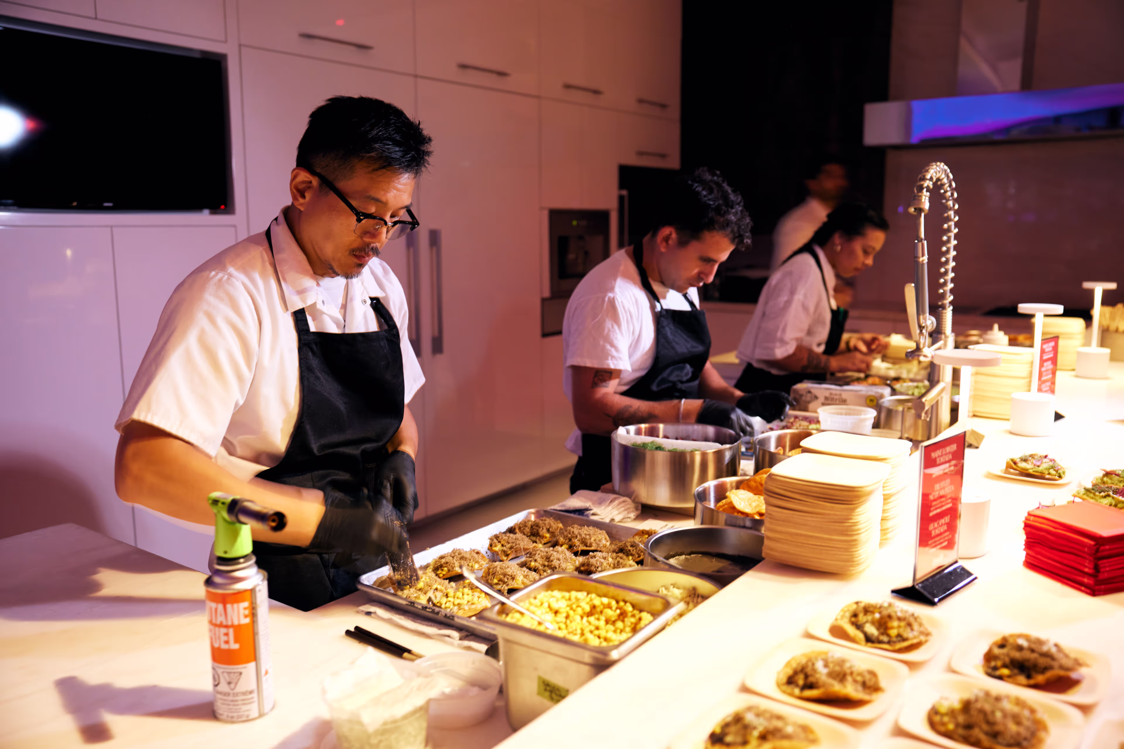 Three chefs in white shirts and black aprons preparing food on plates at a kitchen counter with various ingredients and utensils.