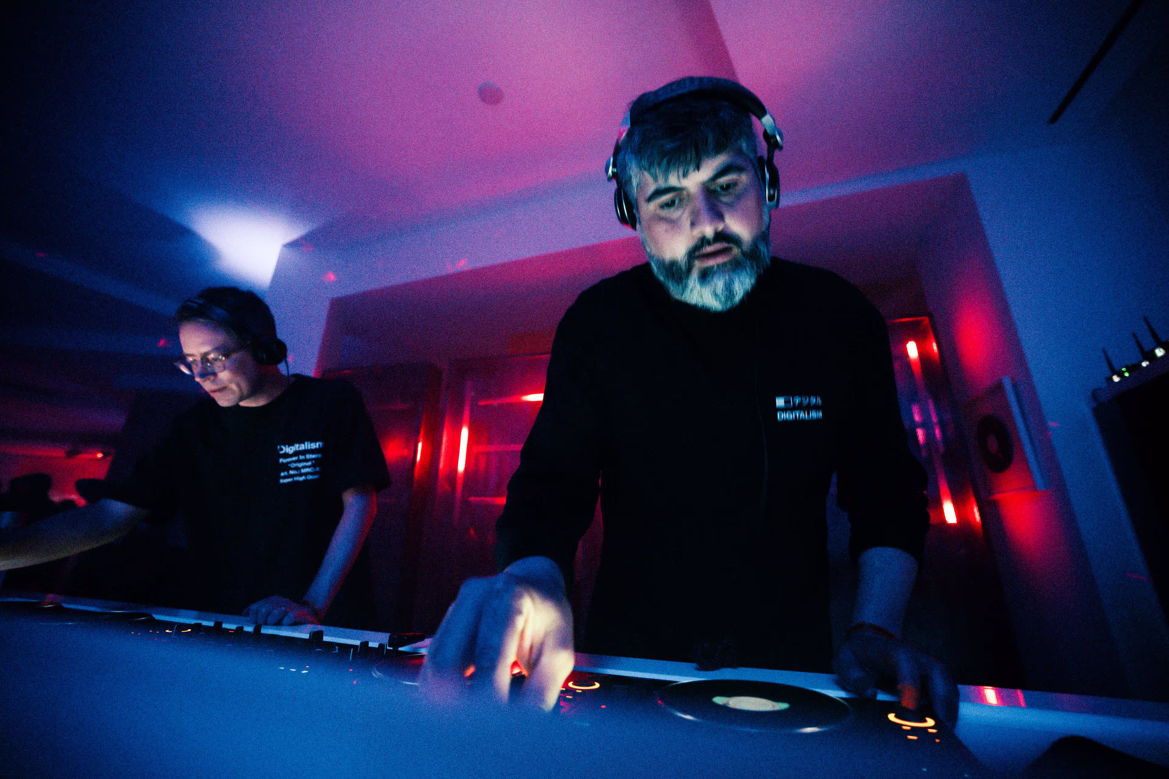 Two DJs wearing headphones performing with mixing equipment under blue and red club lighting.