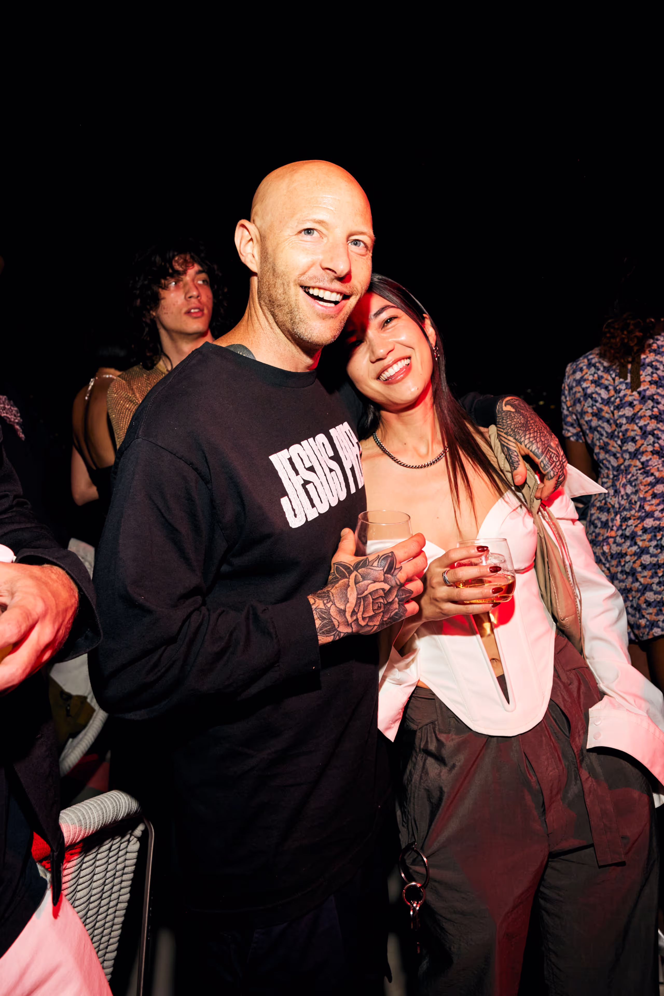 Smiling bald man with rose tattoos on his hand wearing a black sweatshirt is hugging a smiling woman in a white off-shoulder top holding drinks at a party.