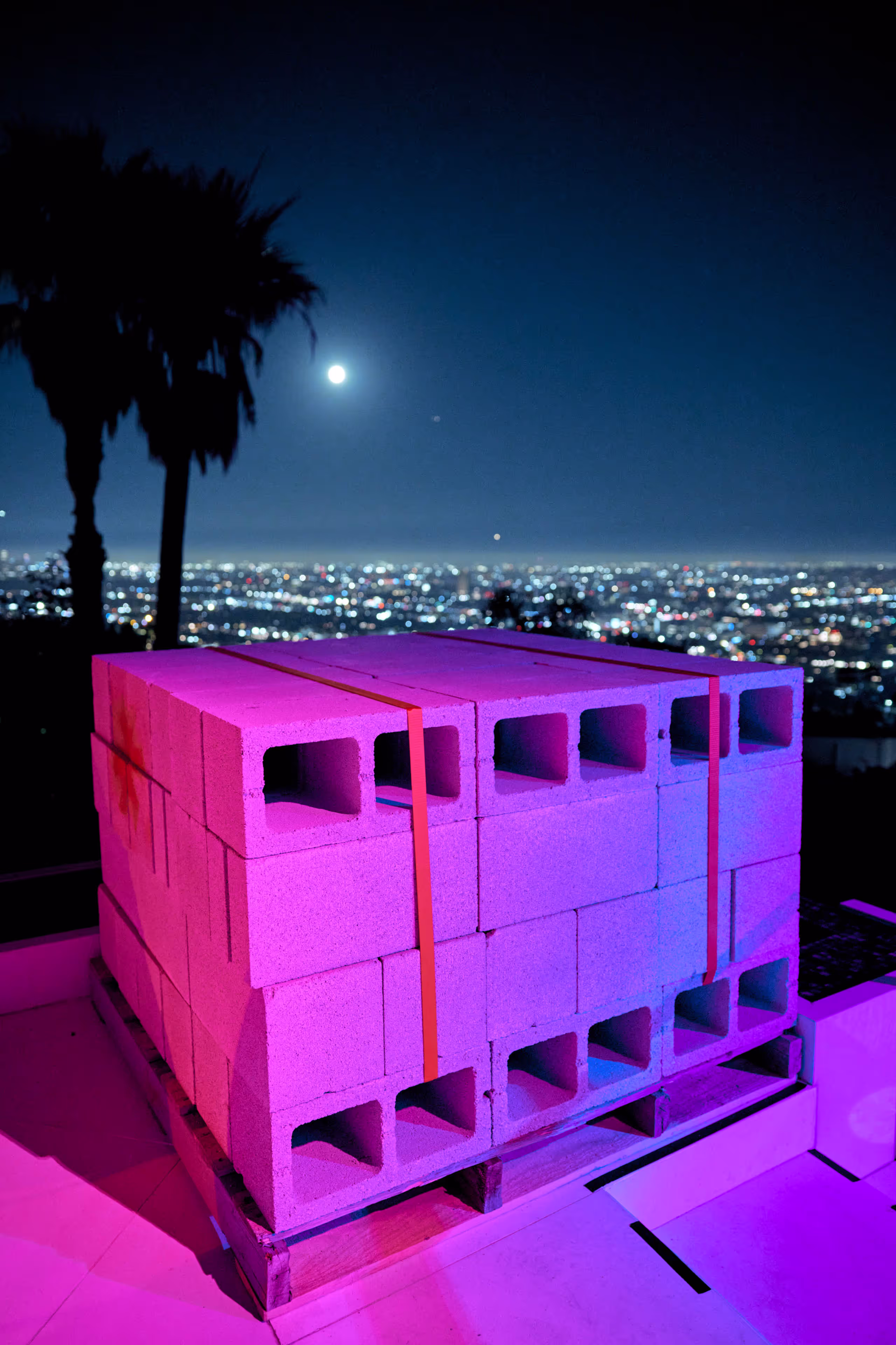 Stack of hollow concrete blocks on a wooden pallet under purple and blue lighting with a cityscape and full moon in the night background.
