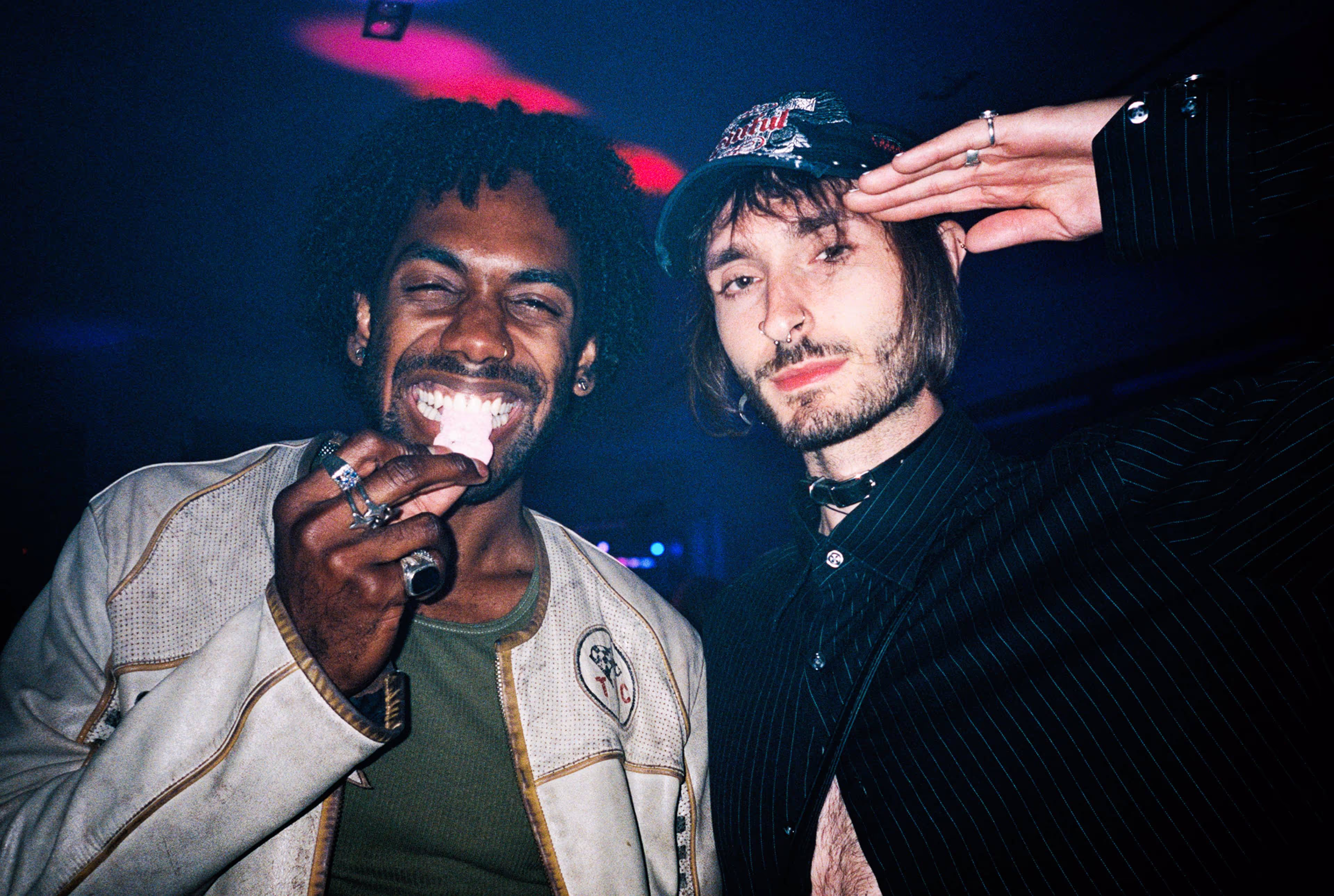 Two young men posing closely together in a dimly lit setting.