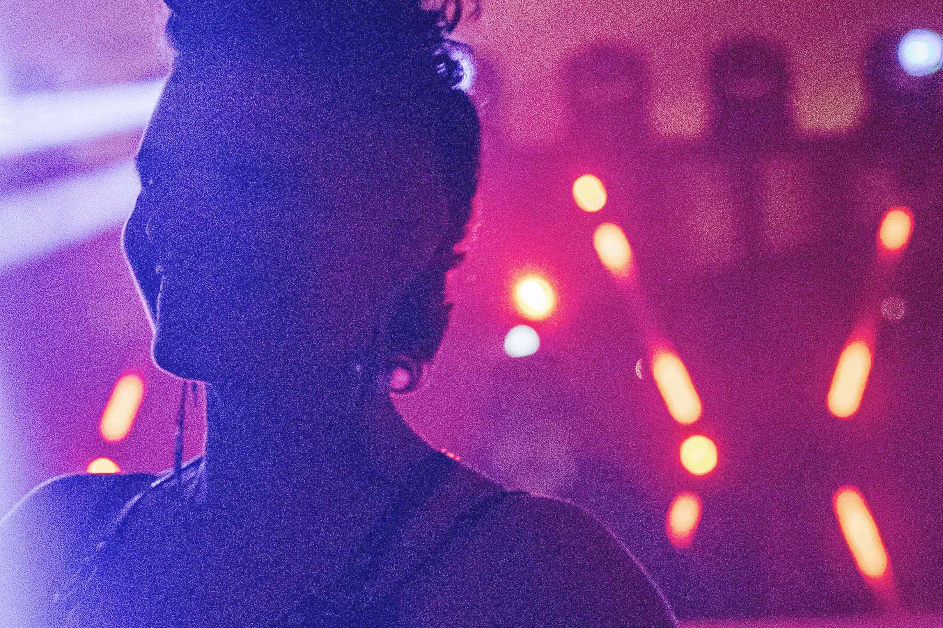 Silhouette of a person with curly hair against a background of blurred colorful stage lights in pink and purple hues.