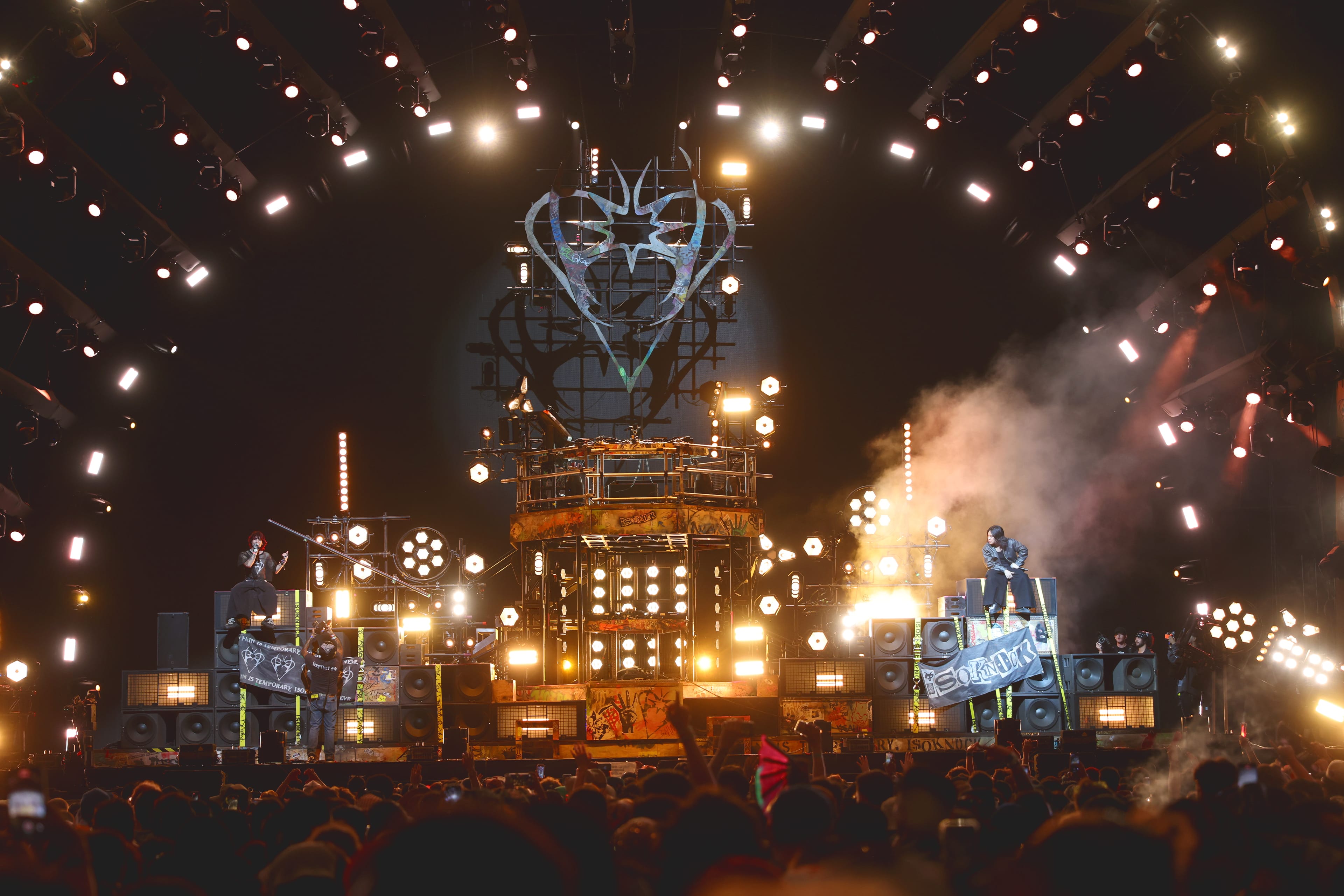 Concert stage with multiple bright lights and performers elevated on speaker stacks.